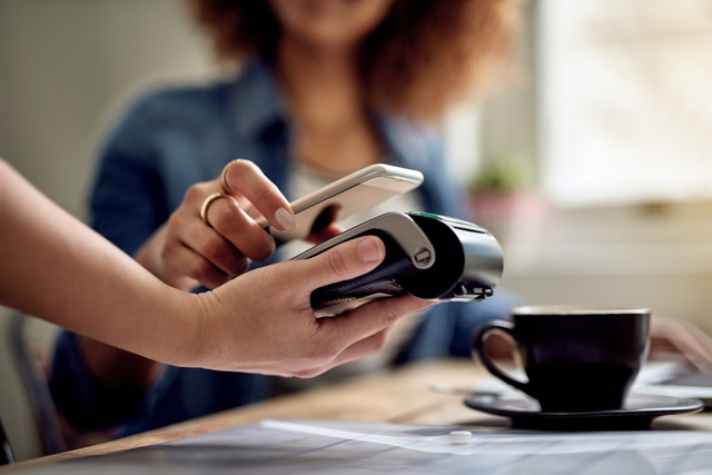 Hand houdt een smartphone tegen een betaalterminal voor een contactloze betaling aan tafel, met een kop koffie in een rustige horecasetting