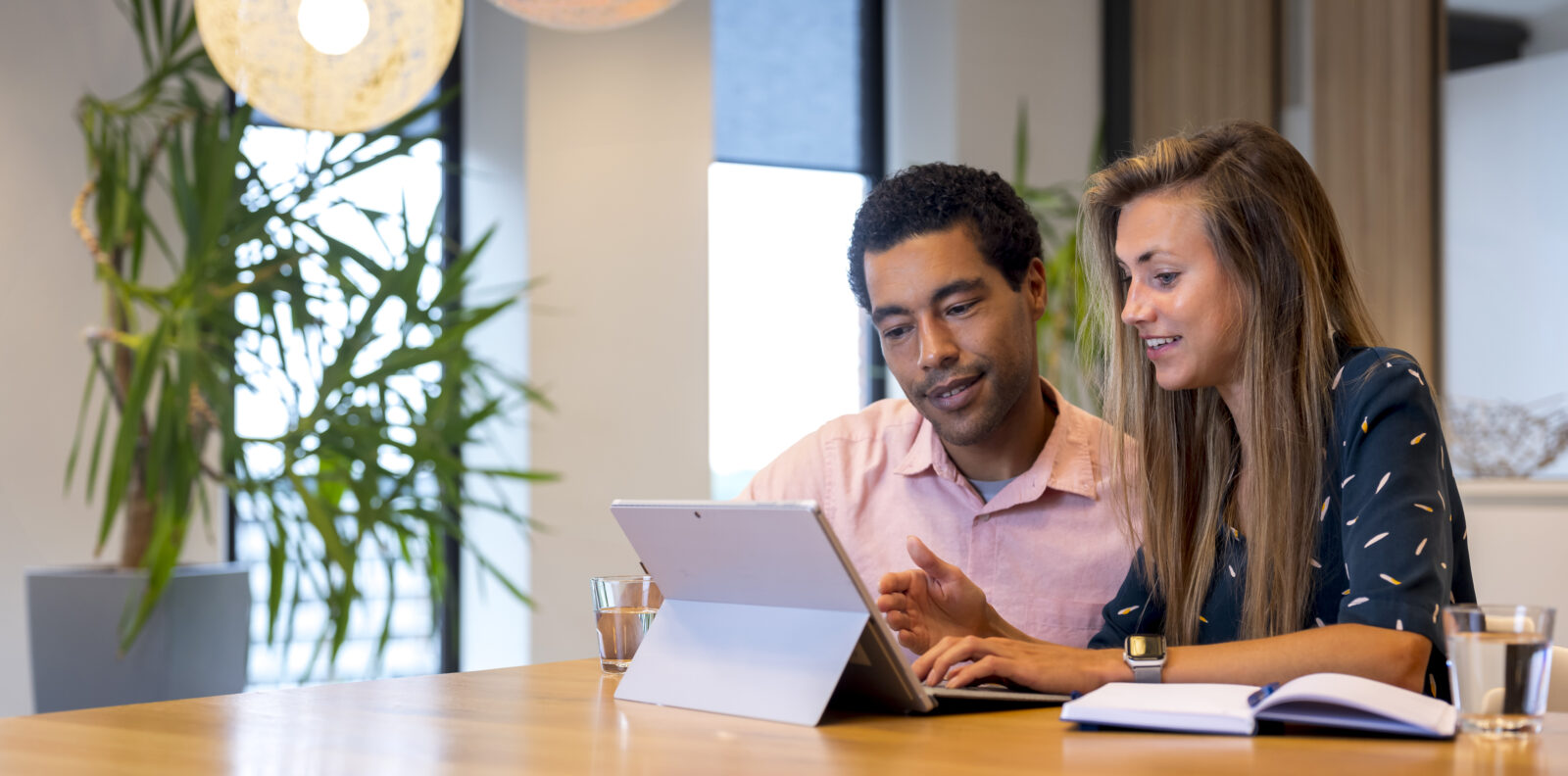 Afbeelding van twee personen die samen werken aan een tablet aan een vergadertafel in een lichte kantooromgeving