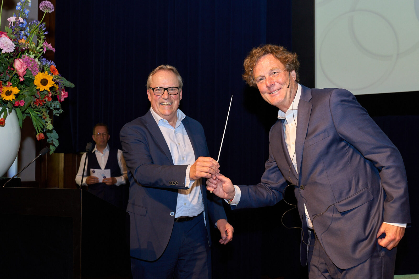 Gijs Boudewijn en Arjan Bol poseren op het podium tijdens het afscheidscongres van Gijs Boudewijn, waarbij Gijs symbolisch een stok overhandigt aan Arjan Bol als overdracht van het stokje als algemeen directeur van Betaalvereniging Nederland
