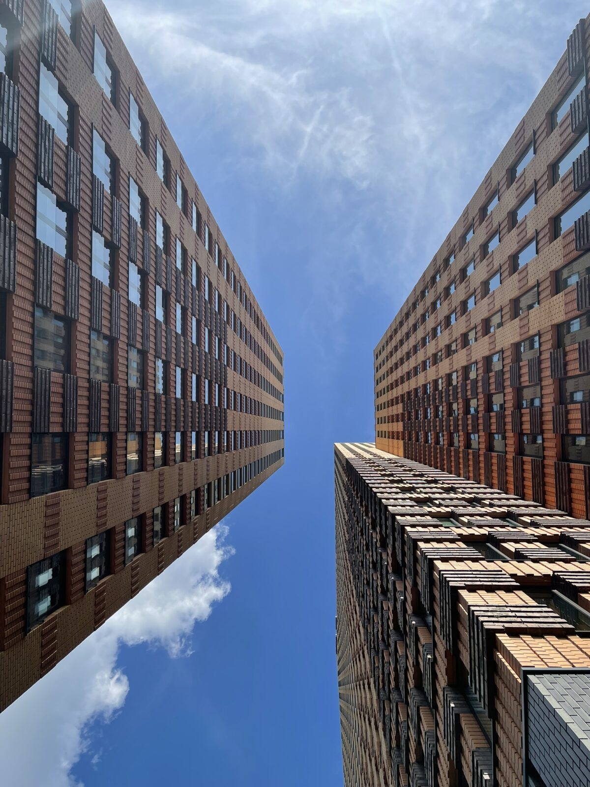 Foto genomen vanuit een laag standpunt tussen twee moderne bakstenen kantoorgebouwen met strakke lijnen, met daarboven een open blauwe lucht