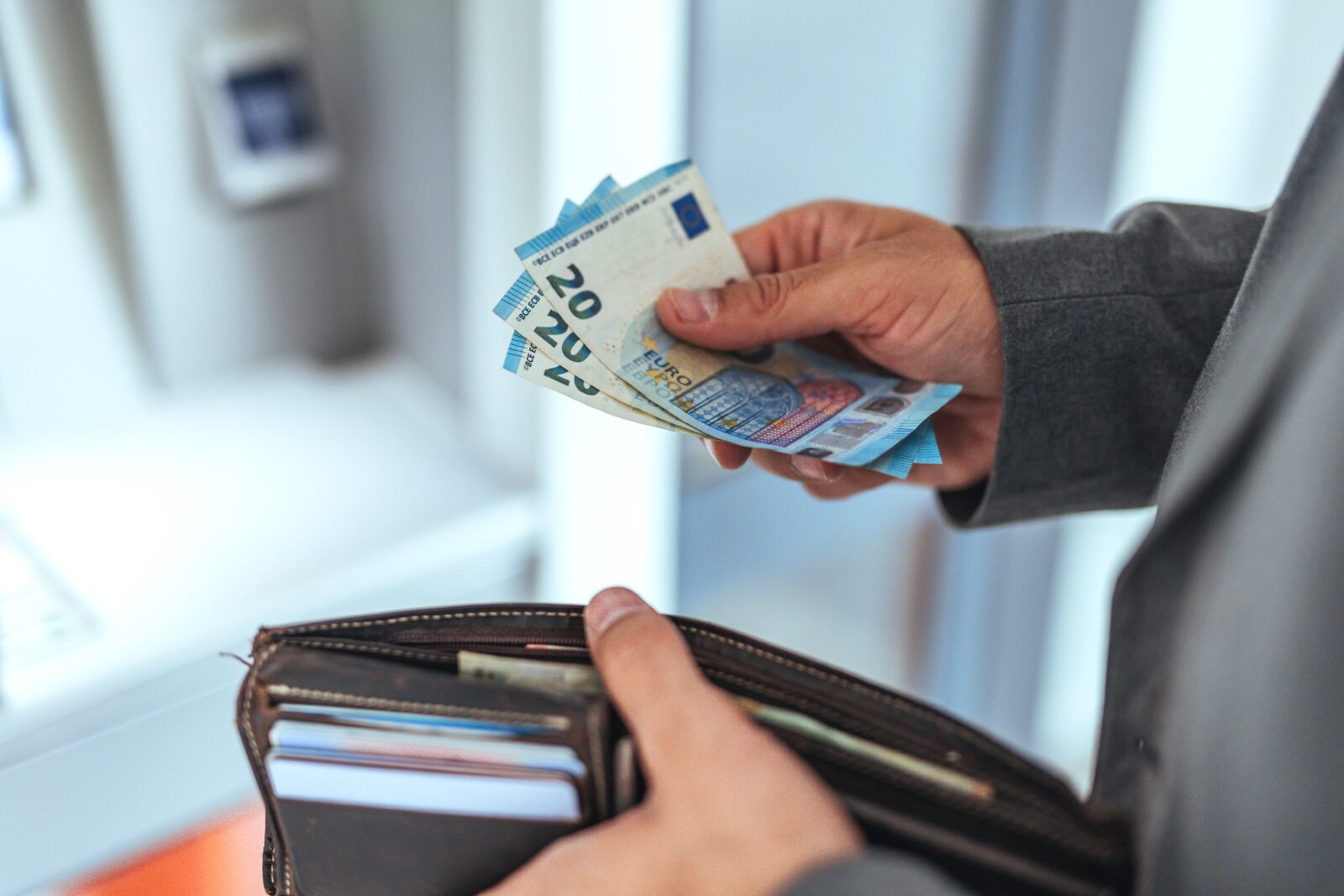 Afbeelding van een persoon voor een geldautomaat, met in de rechterhand drie biljetten van twintig euro en in de linkerhand een bruine portemonnee met pasjes